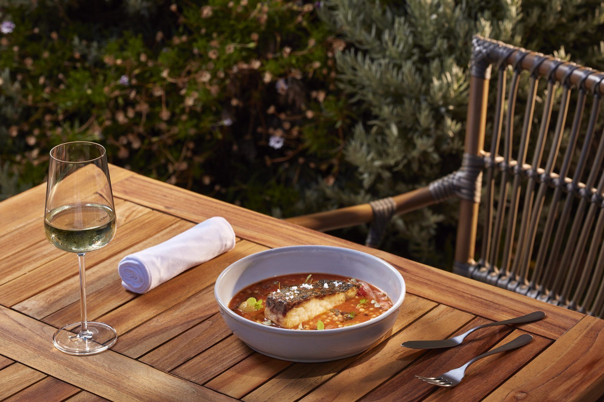 Una mesa de madera con un tazón de sopa y una copa de vino blanco, una servilleta doblada y cubiertos.