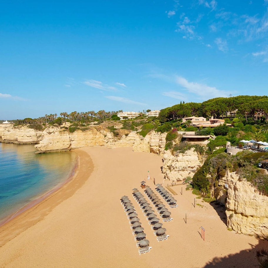 A beach with a row of sun loungers and umbrellas, a rocky coastline, and buildings in the background.