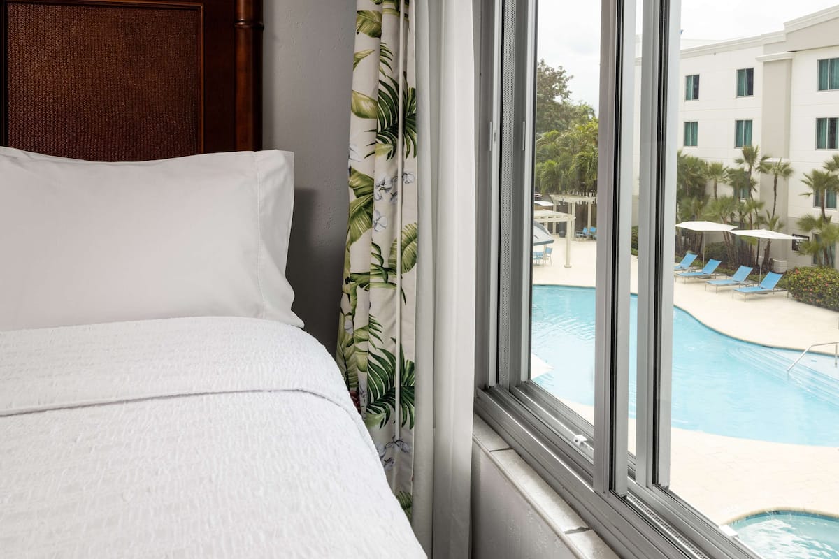 A hotel room with a bed, a window with a view of a swimming pool and lounge chairs.
