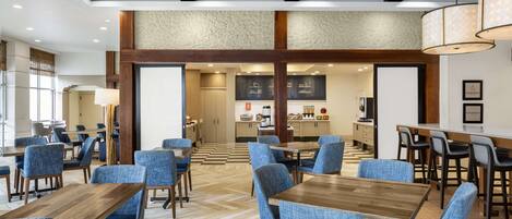 Un hall d’hôtel doté de plusieurs espaces salon, une cuisine à l’arrière-plan et un plafond orné de motifs.