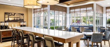 A modern hotel lobby with a long bar area, seating, and large windows offering views of the outdoors.