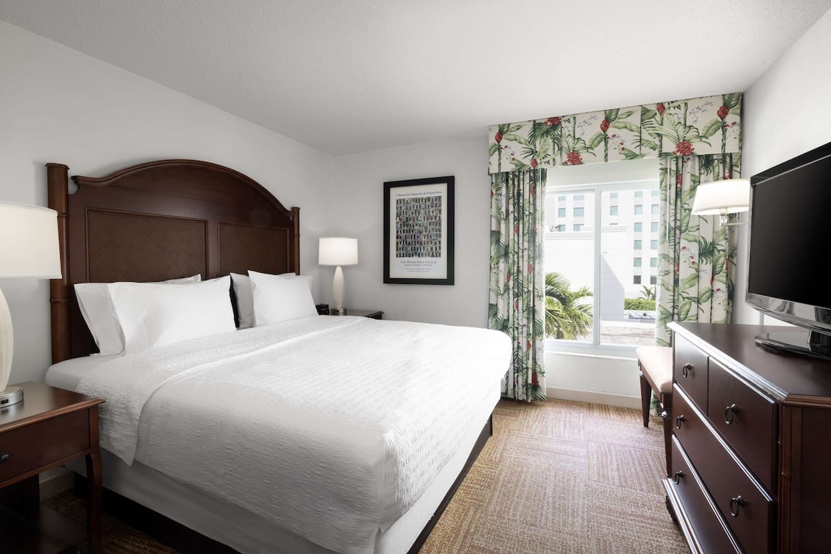 A hotel room with a large bed, a TV, a nightstand, a dresser, and a window with curtains.