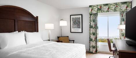 A hotel room with a large bed, two bedside lamps, a desk with a computer, a chair, and a window with curtains.