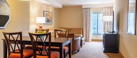 A hotel room with a dining area, a sofa, a TV, and a window with curtains.