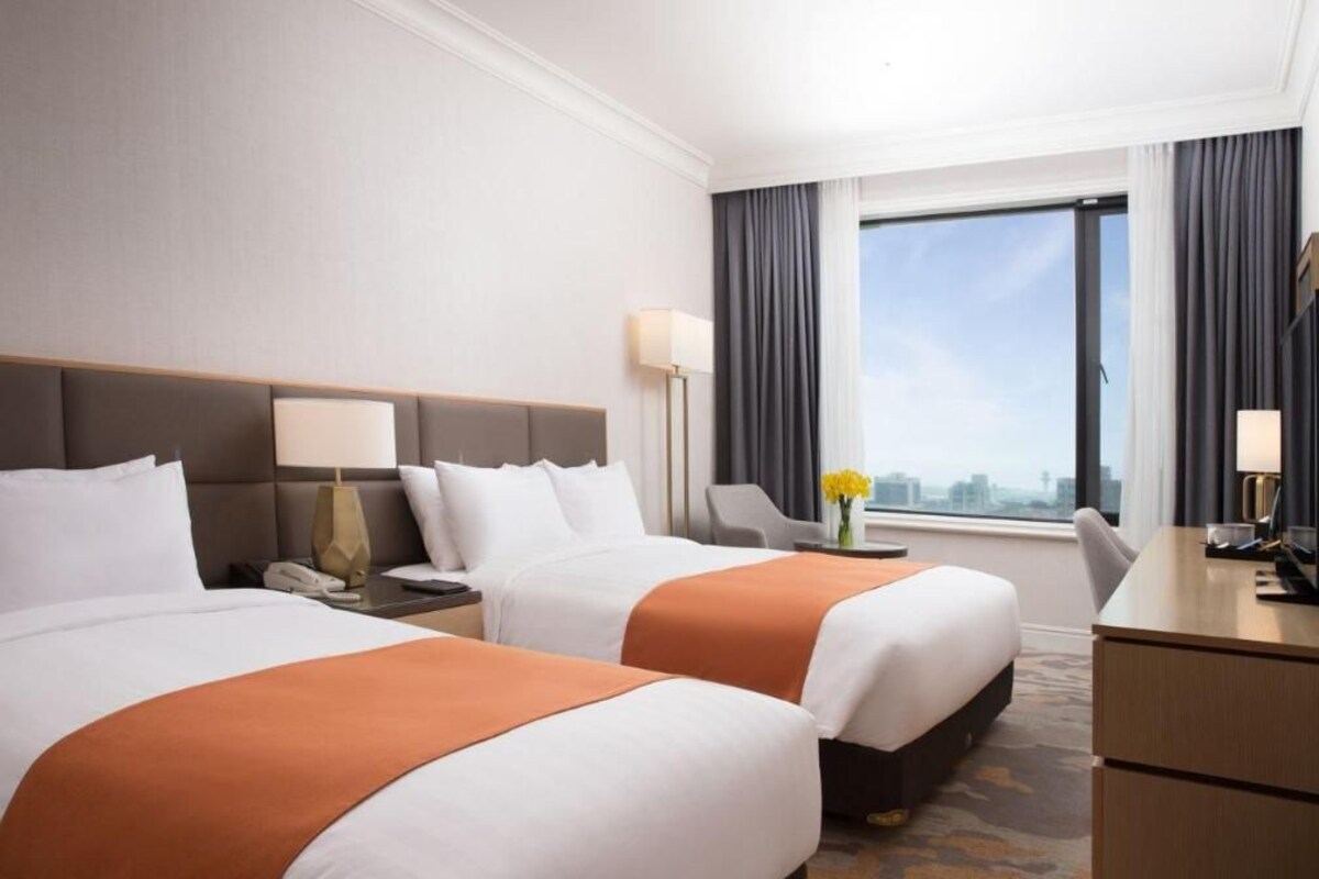 Une chambre d’hôtel avec deux lits, un bureau, une chaise, une lampe et une vue sur la ville par la fenêtre.