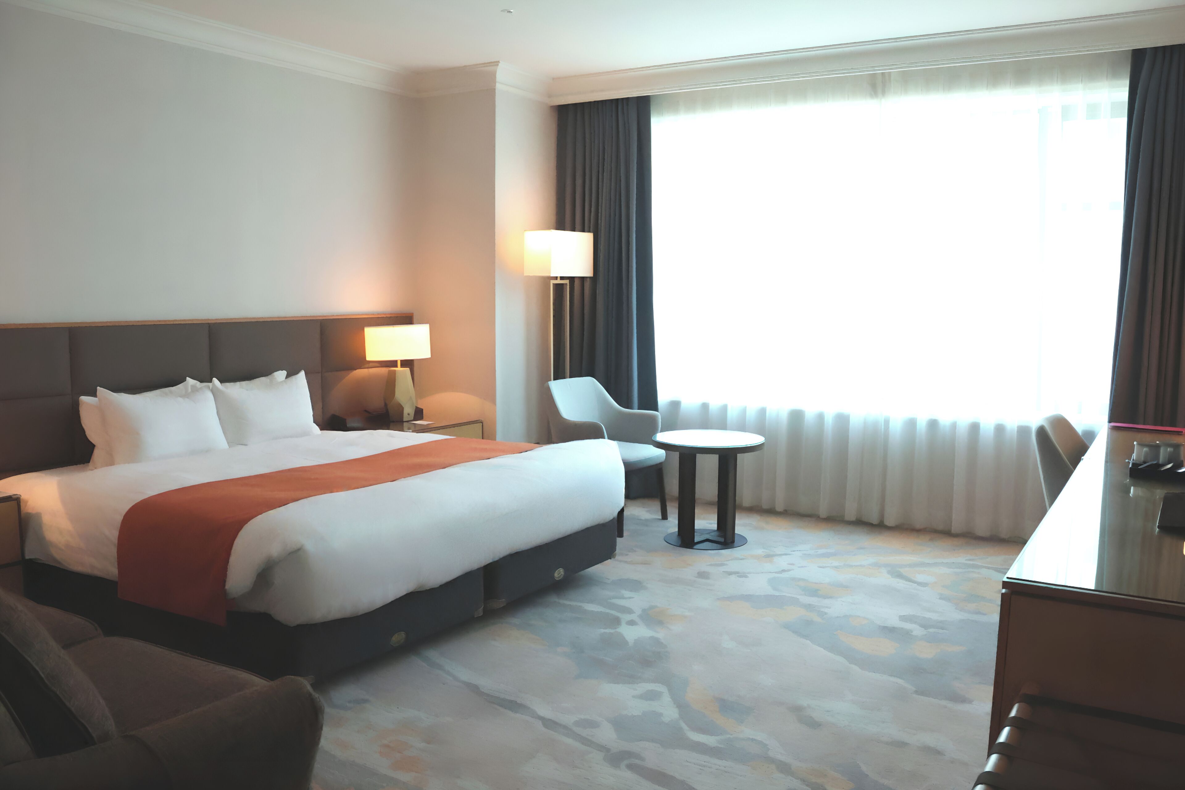 A hotel room with a large bed, a desk, a chair, a lamp, and a view of the outdoors.
