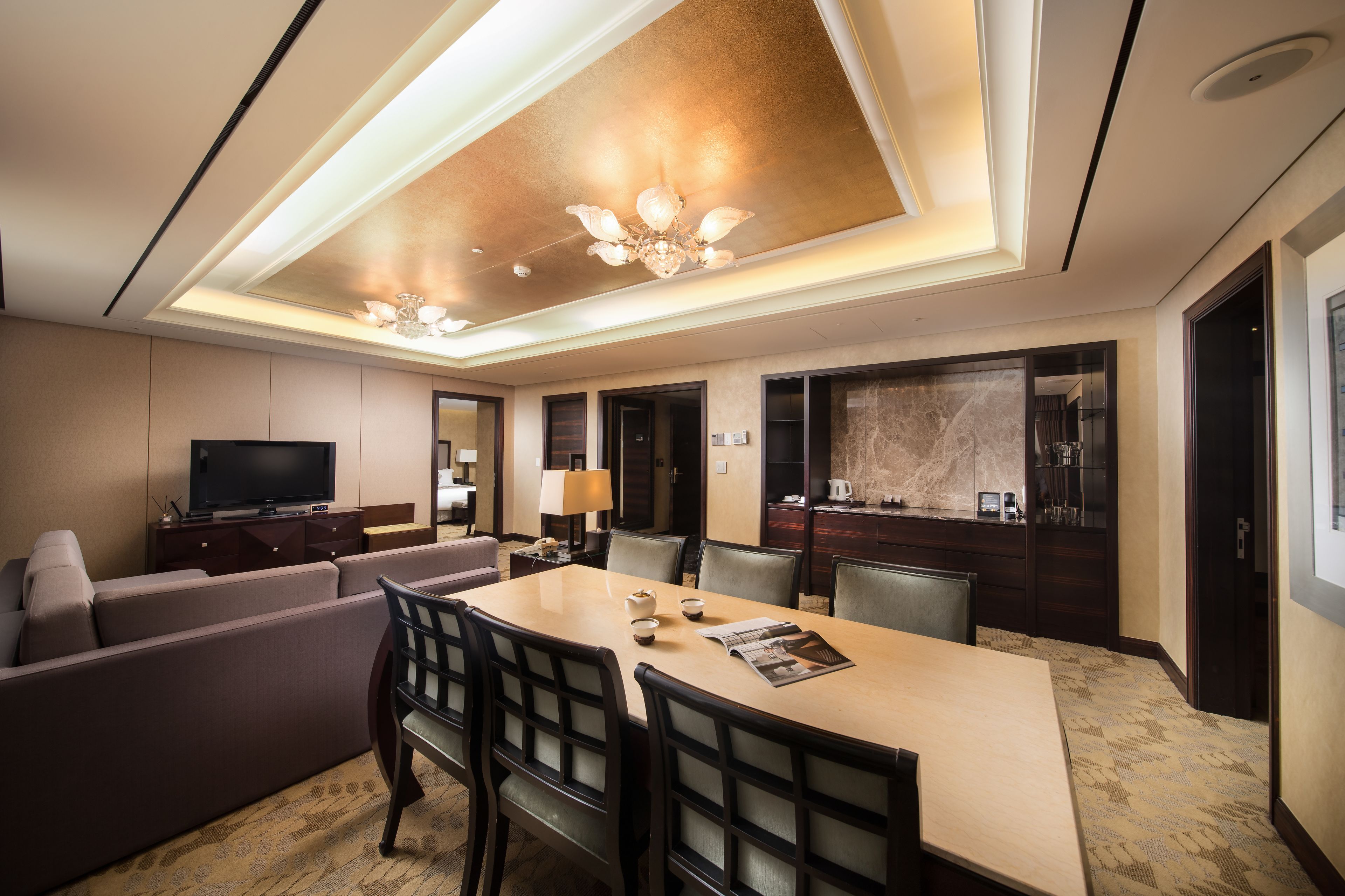 A modern hotel room with a dining area, a sofa, a television, and a kitchen area.
