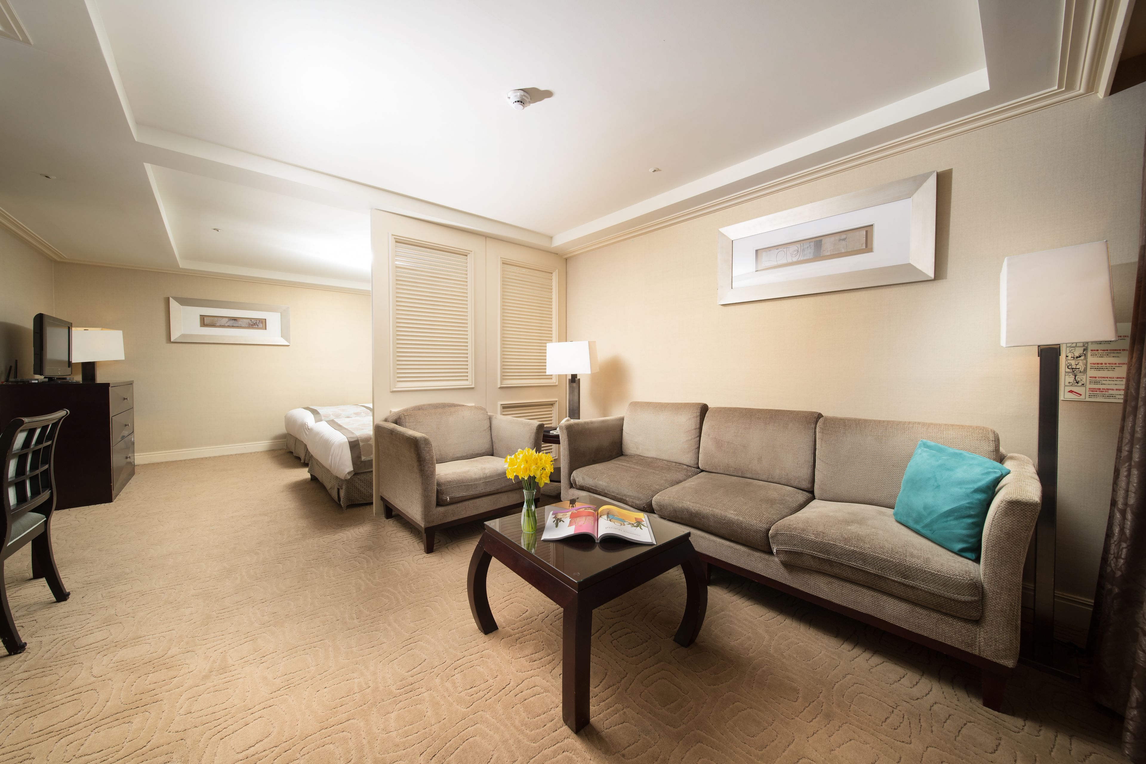 Un salón con alfombra color beige, un sofá marrón, un sillón marrón, una mesa de centro de madera con un libro y flores, y un televisor sobre un mueble.