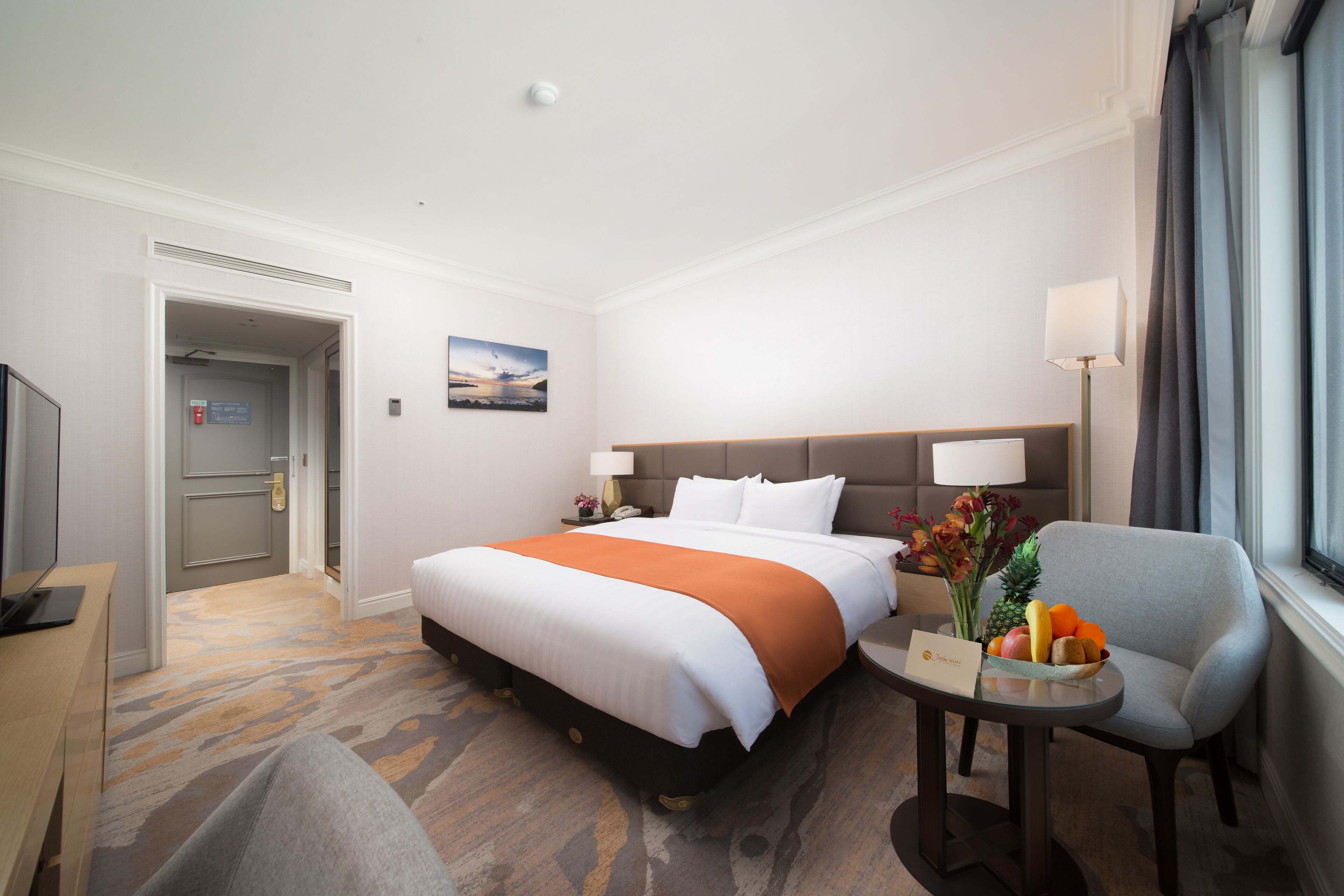 Ein Hotelzimmer mit Bett, Schreibtisch, Stuhl, einem kleinen Tisch mit Obst und einem Fenster mit Vorhängen.
