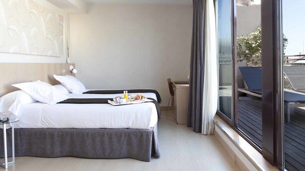 A hotel room with a bed, a small table with a tray of breakfast, a chair, and a large window with a view of the outside. - Hotel Ópera (Madrid)