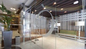 A modern reception area with a glass partition, a green bench, and a potted plant. - Hotel Ópera (Madrid)