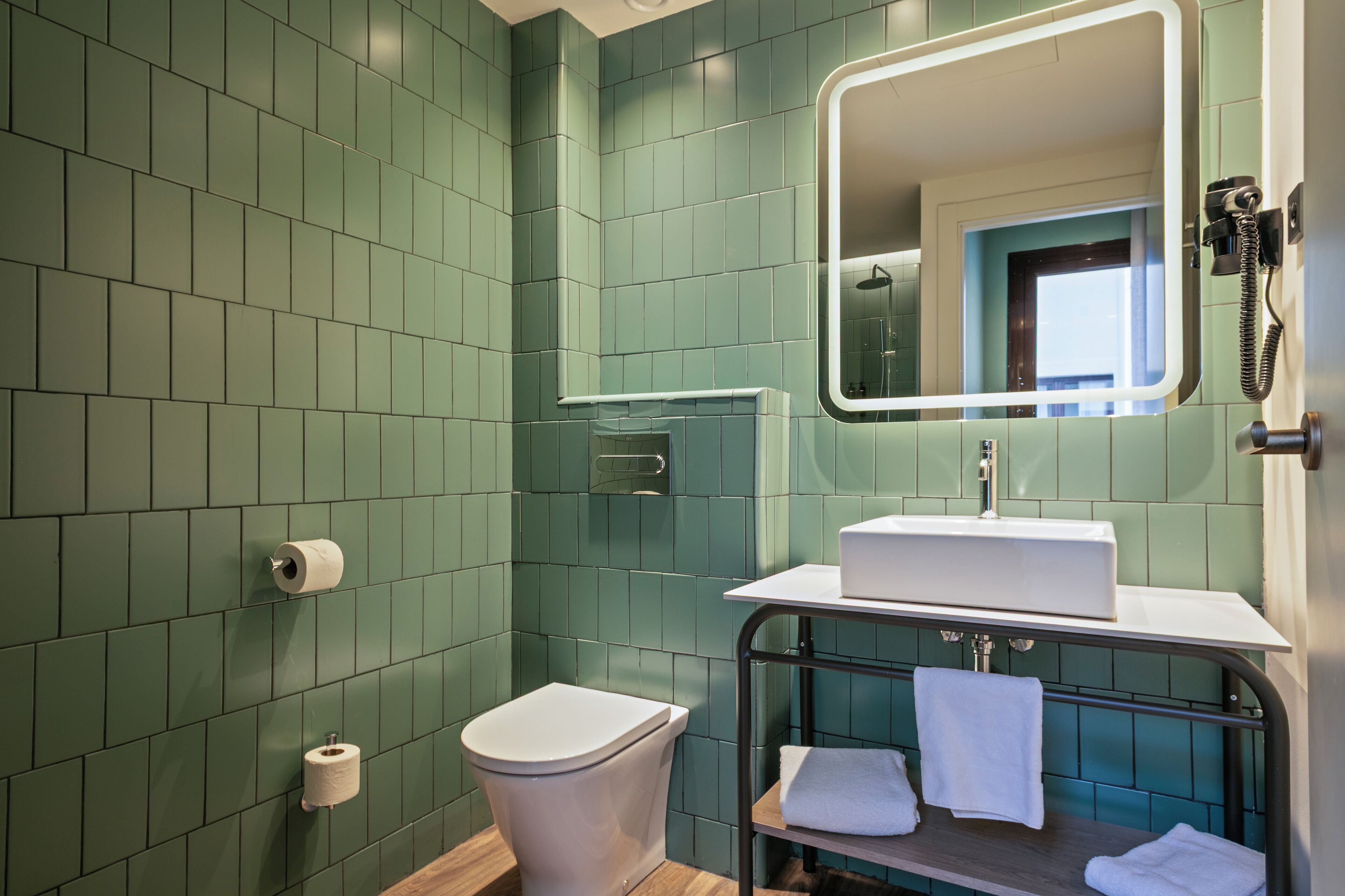 Un bany amb rajoles verdes, un vàter blanc, un lavabo amb mirall i un suport per a tovalloles.