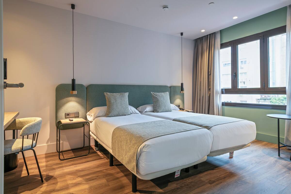 Une chambre d’hôtel avec deux lits, un bureau, une chaise, une petite table et une fenêtre avec des rideaux.