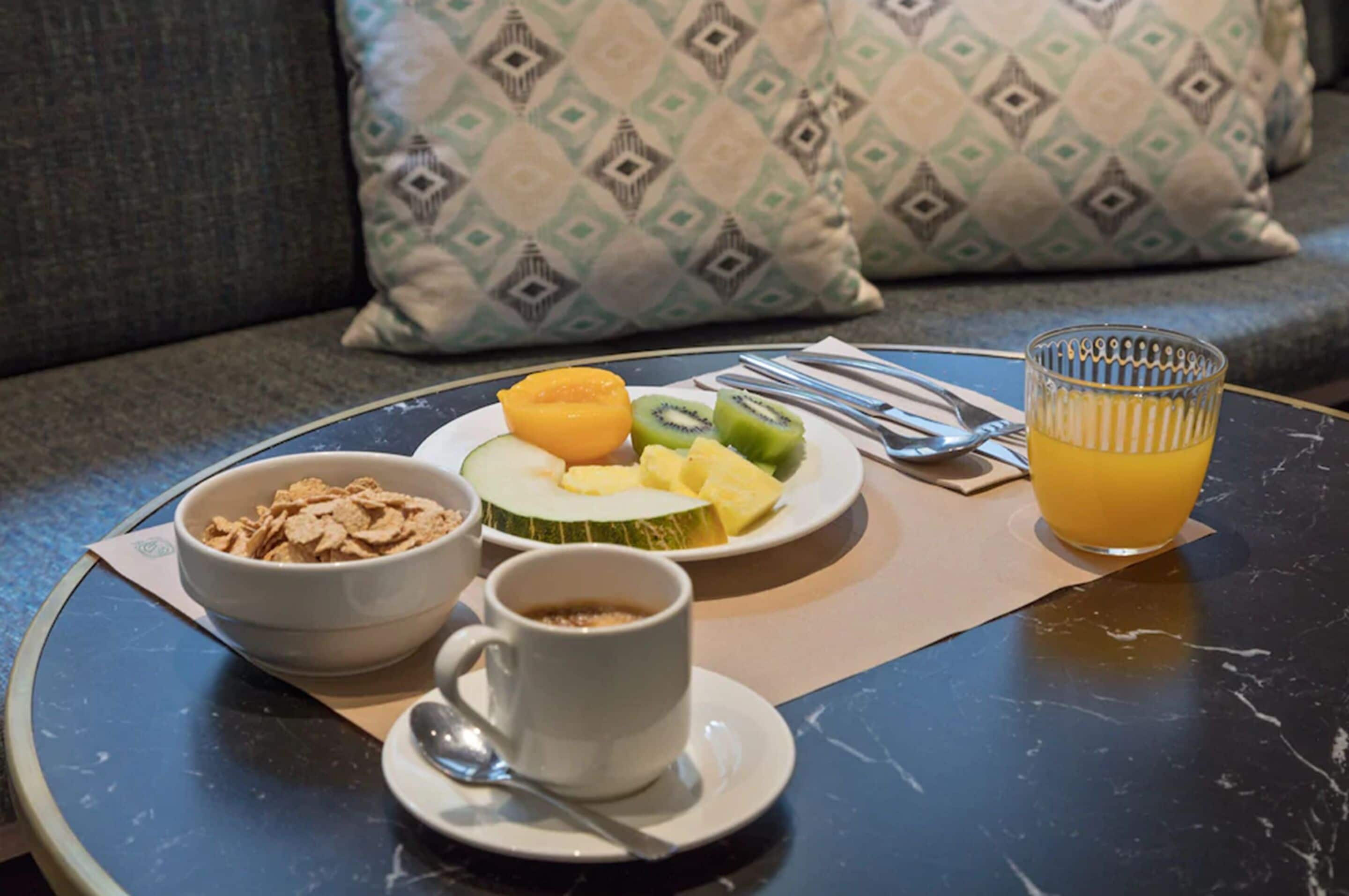A breakfast setup on a round table with a bowl of cereal, a cup of coffee, a plate with fruit, and a glass of orange juice.