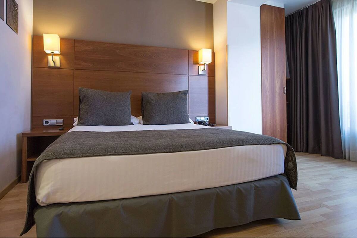 Une chambre d’hôtel avec un grand lit, deux lampes de chevet, une tête de lit en bois et une vue sur un rideau.