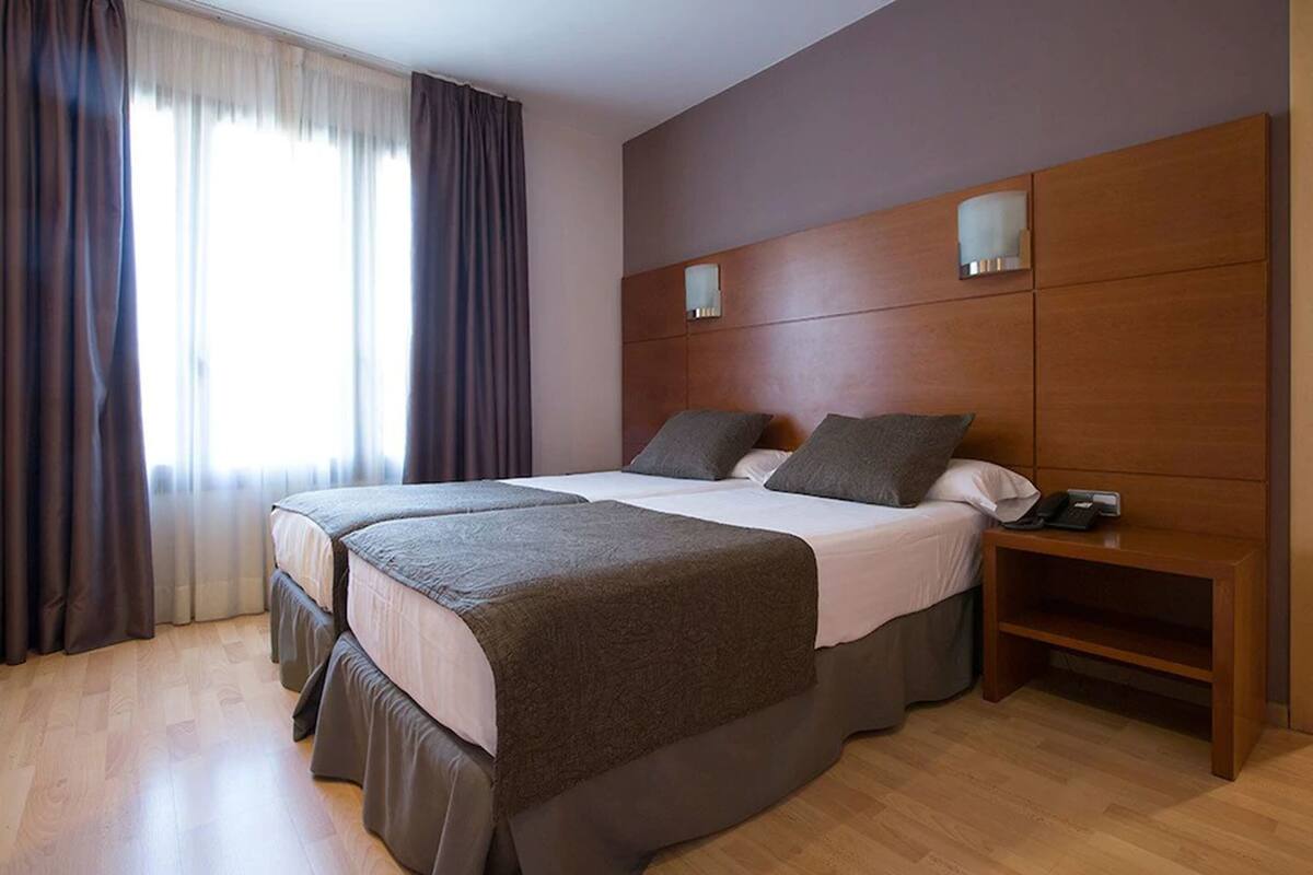 Une chambre d’hôtel avec deux lits, une tête de lit en bois, une table de chevet avec un téléphone et une fenêtre avec des rideaux.