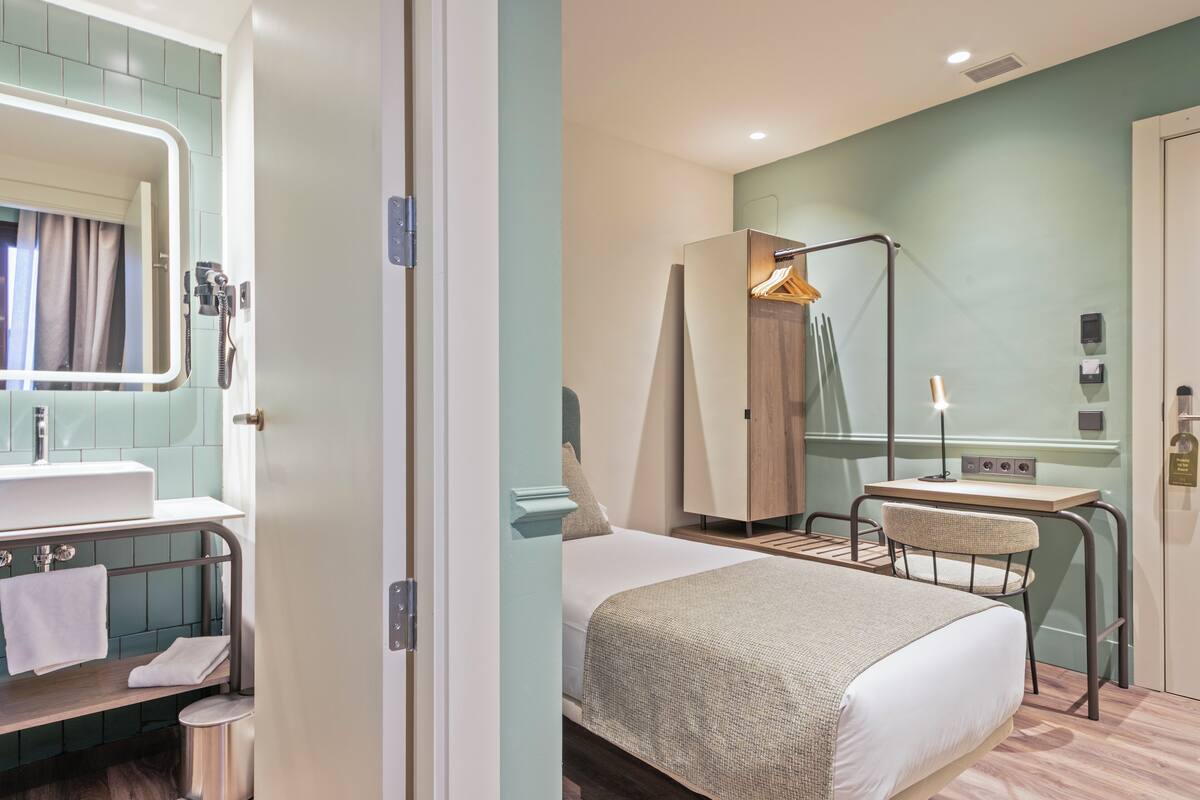 Une chambre d’hôtel équipée d’un lit, d’un bureau, d’une chaise, d’un miroir, d’un lavabo et d’un porte-serviettes.