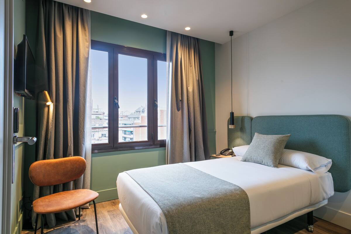 Une chambre d’hôtel avec un lit, une chaise, un bureau et une fenêtre avec des rideaux.