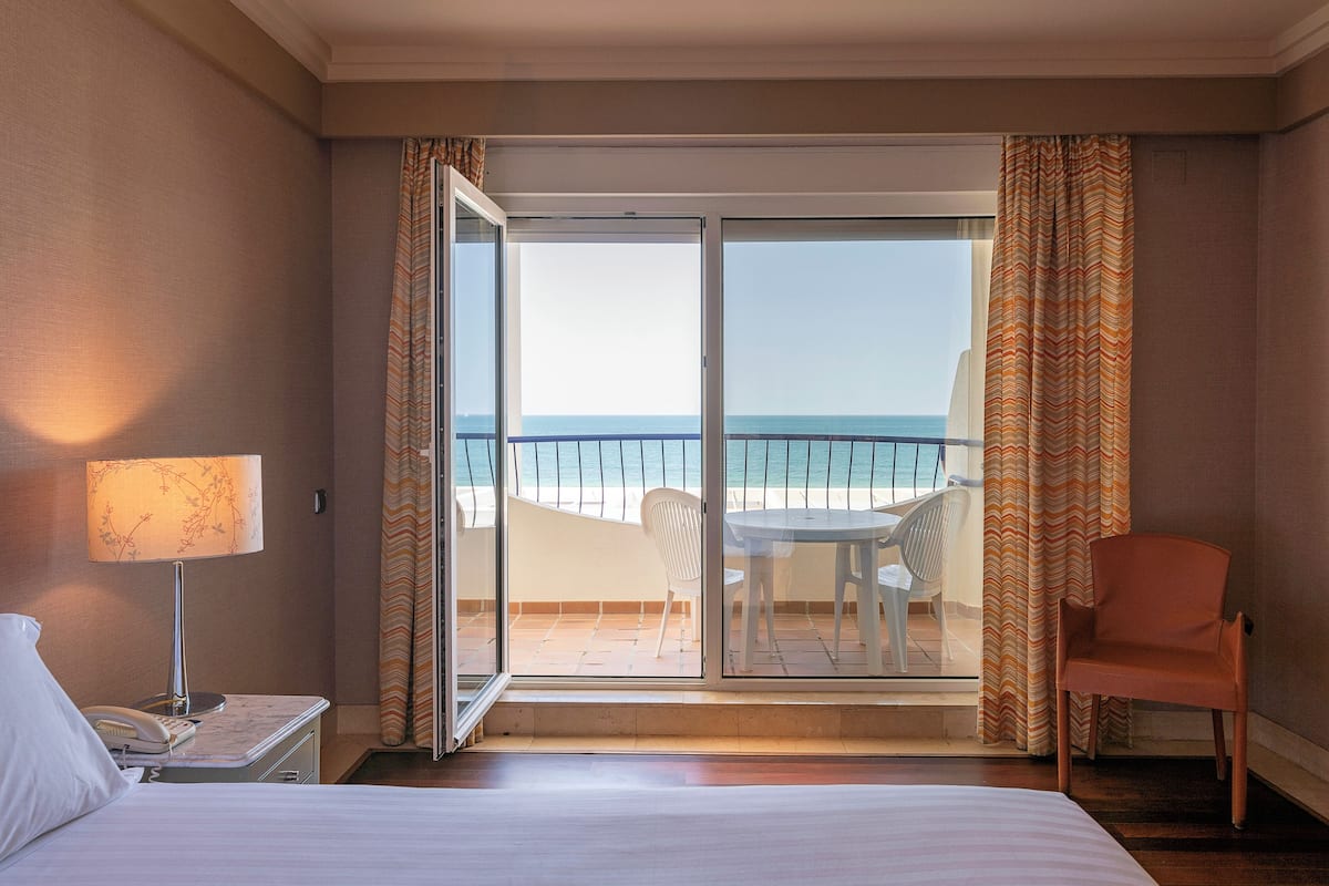 Habitación de hotel con cama, mesita de noche con lámpara, silla y vista al océano a través de una puerta corrediza de vidrio.
