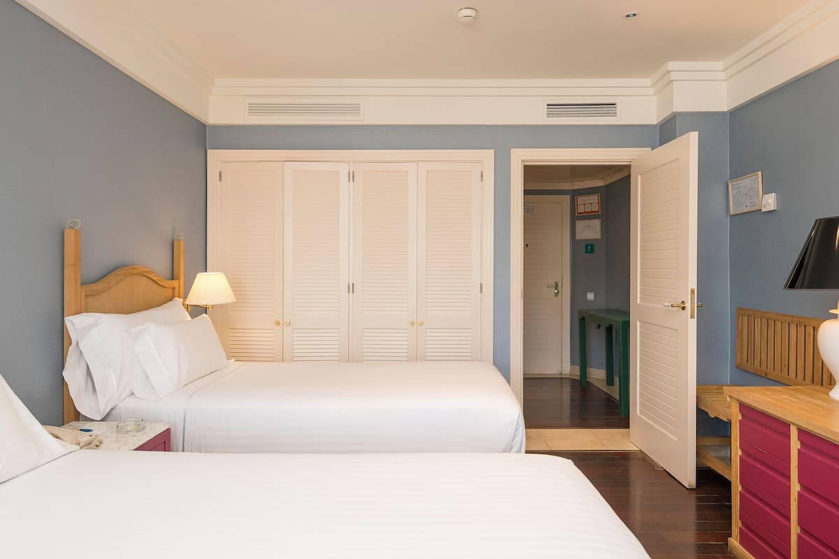 Habitación de hotel con dos camas, cabecera de madera, lámpara de noche, teléfono y cómoda roja.