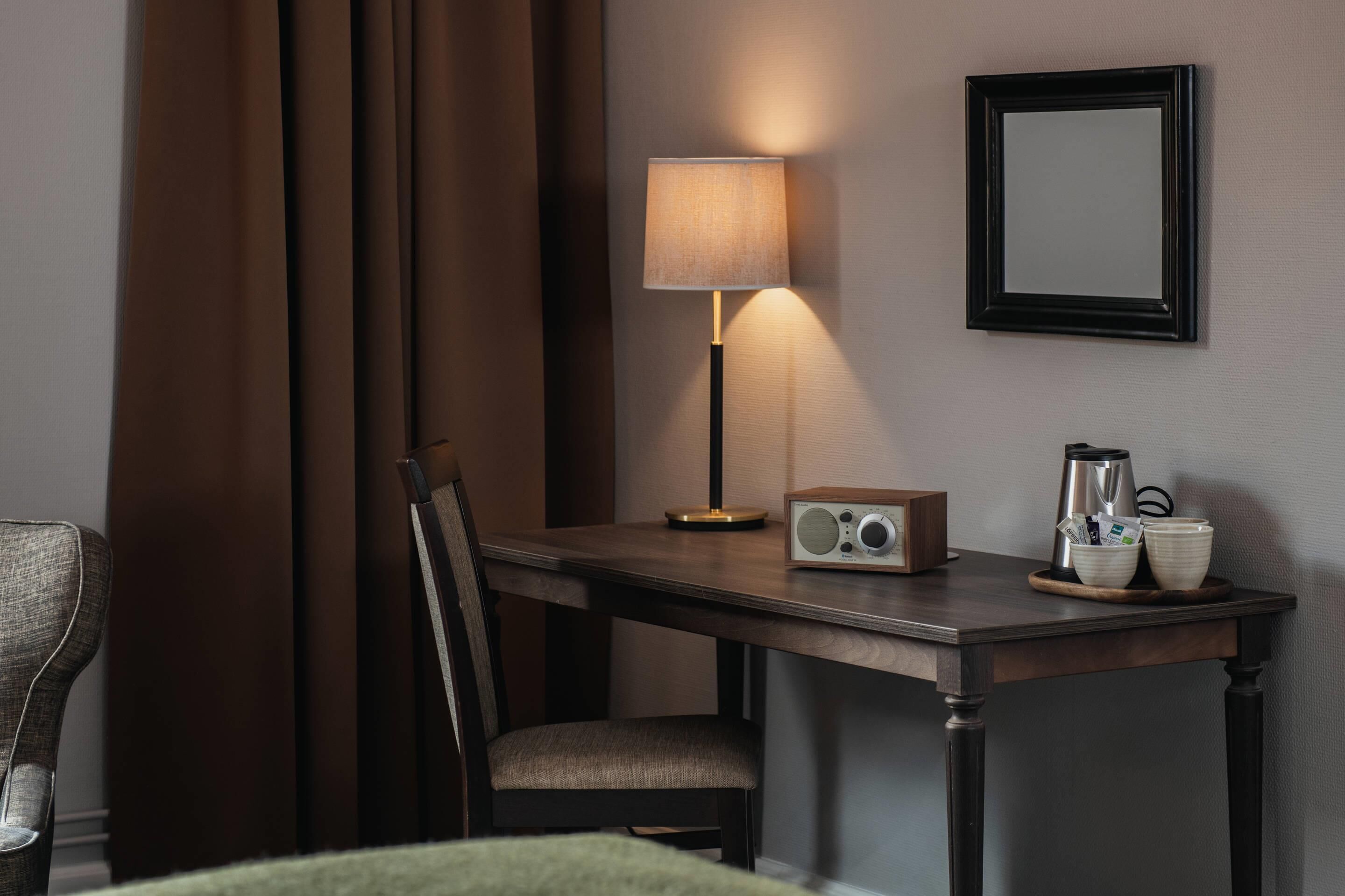 A hotel room with a wooden desk, a chair, a table lamp, a radio, and a coffee maker.