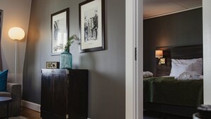 A hotel room with a dark grey wall, a framed picture, a small cabinet with a vase, a bed with a green bedspread, and a nightstand with a lamp. - Home Hotel Uman (Umea)