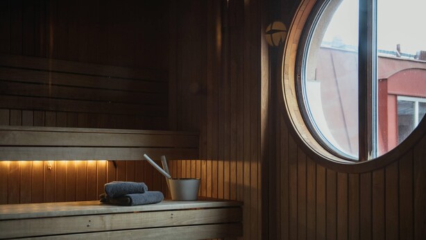 A sauna room with wooden benches, a small table with towels and a bowl, and a window with a view of buildings. - Home Hotel Uman (Umea)