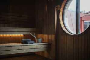 Una sauna amb seients de fusta, una taula petita amb tovellons i un bol, i una finestra que mostra edificis.