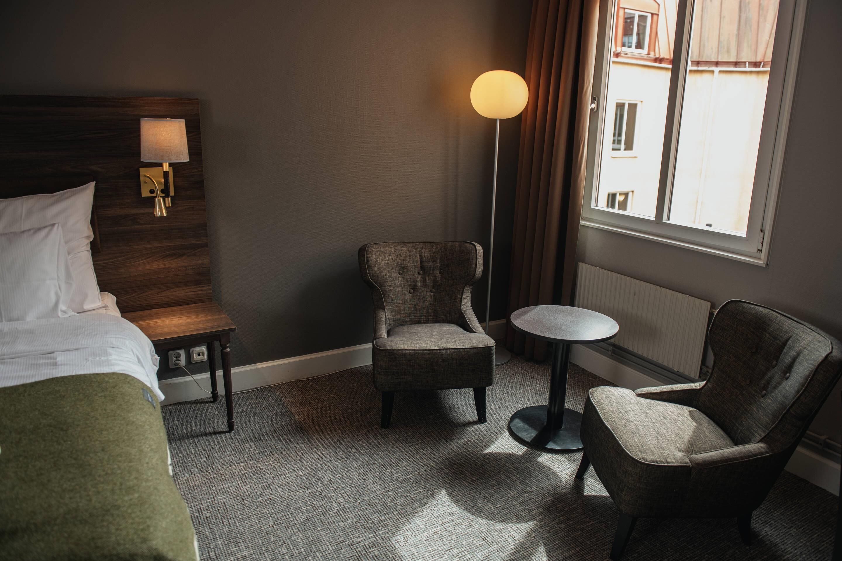 A hotel room with a bed, two armchairs, a small round table, a floor lamp, and a window with a view of buildings.