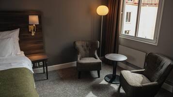 A hotel room with a bed, two armchairs, a small round table, a floor lamp, and a window with a view of buildings.