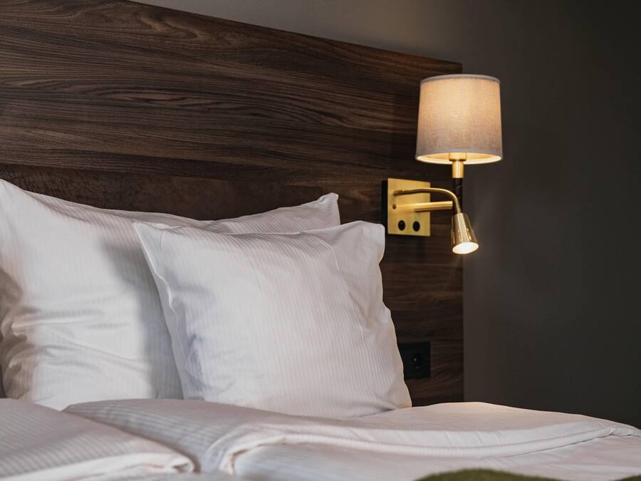 Una cama bien hecha con sábanas blancas, un cabecero de madera y una lámpara fijada a la pared.