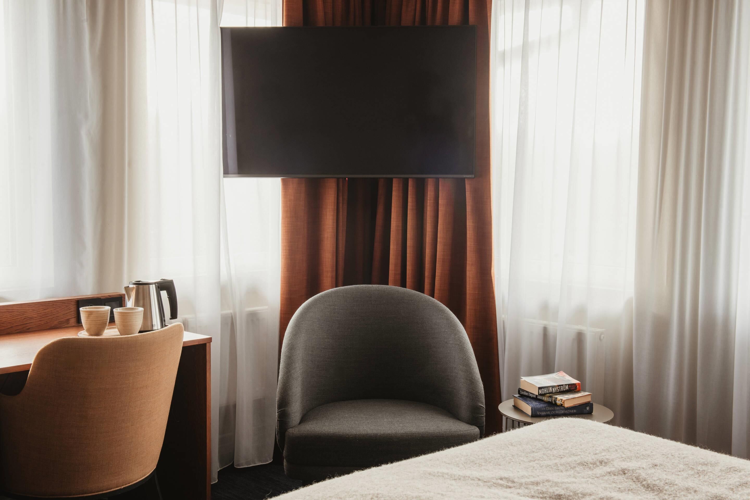 A hotel room with a bed, a desk with a chair, a television, and a small table with books.