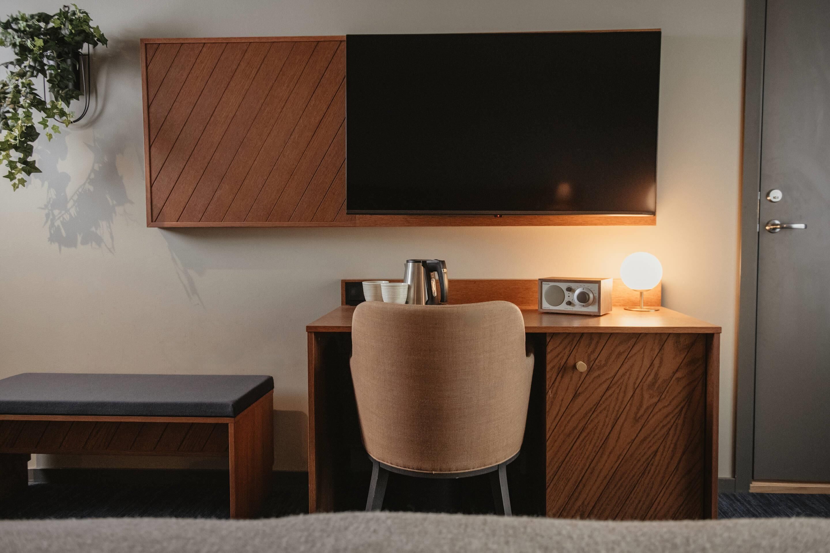 A modern room with a wooden desk, a chair, a flat-screen TV, a radio, and a bedside table.