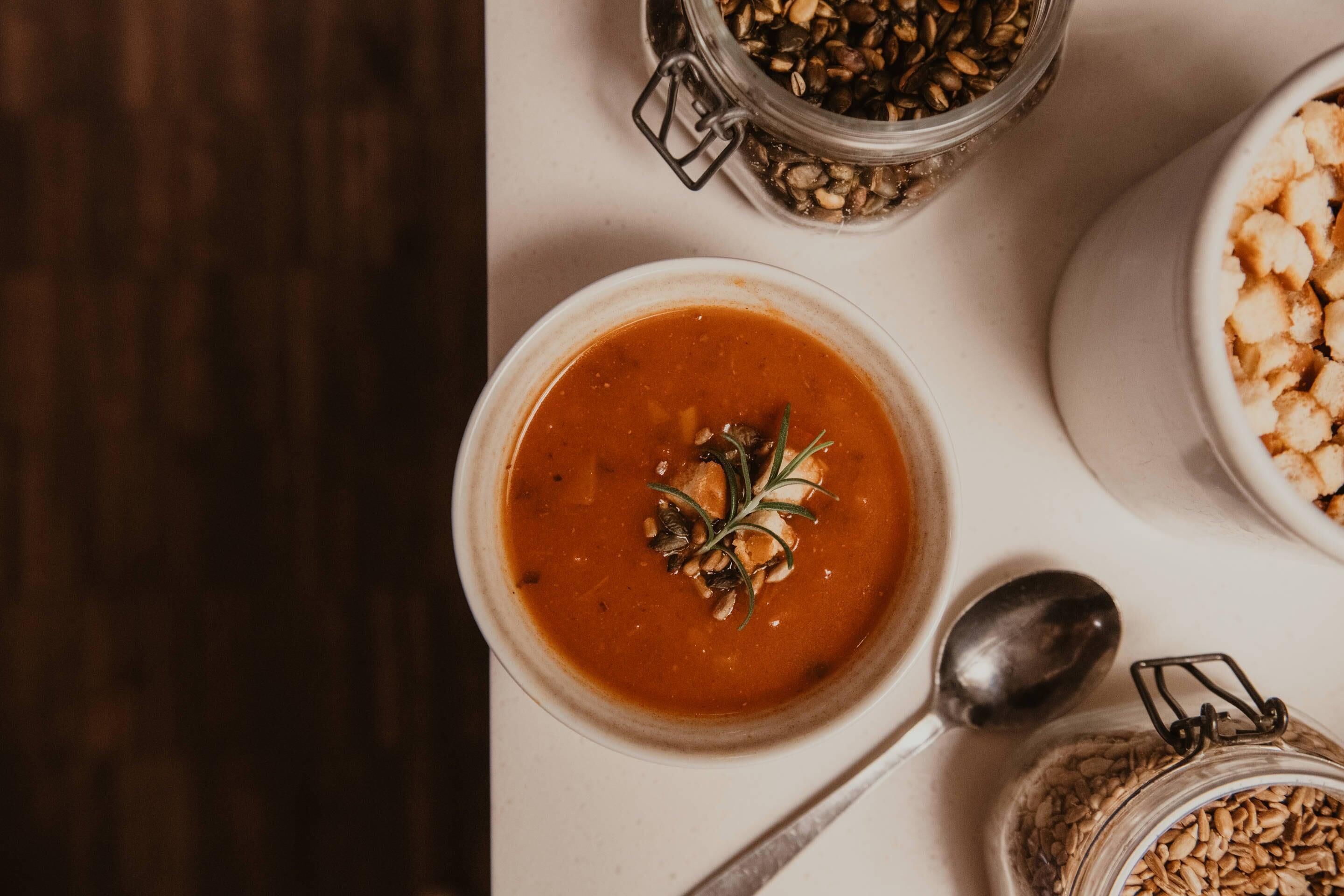 Eine Schüssel Suppe mit einem Rosmarinzweig obenauf, auf einer weißen Oberfläche, neben der ein Löffel liegt.