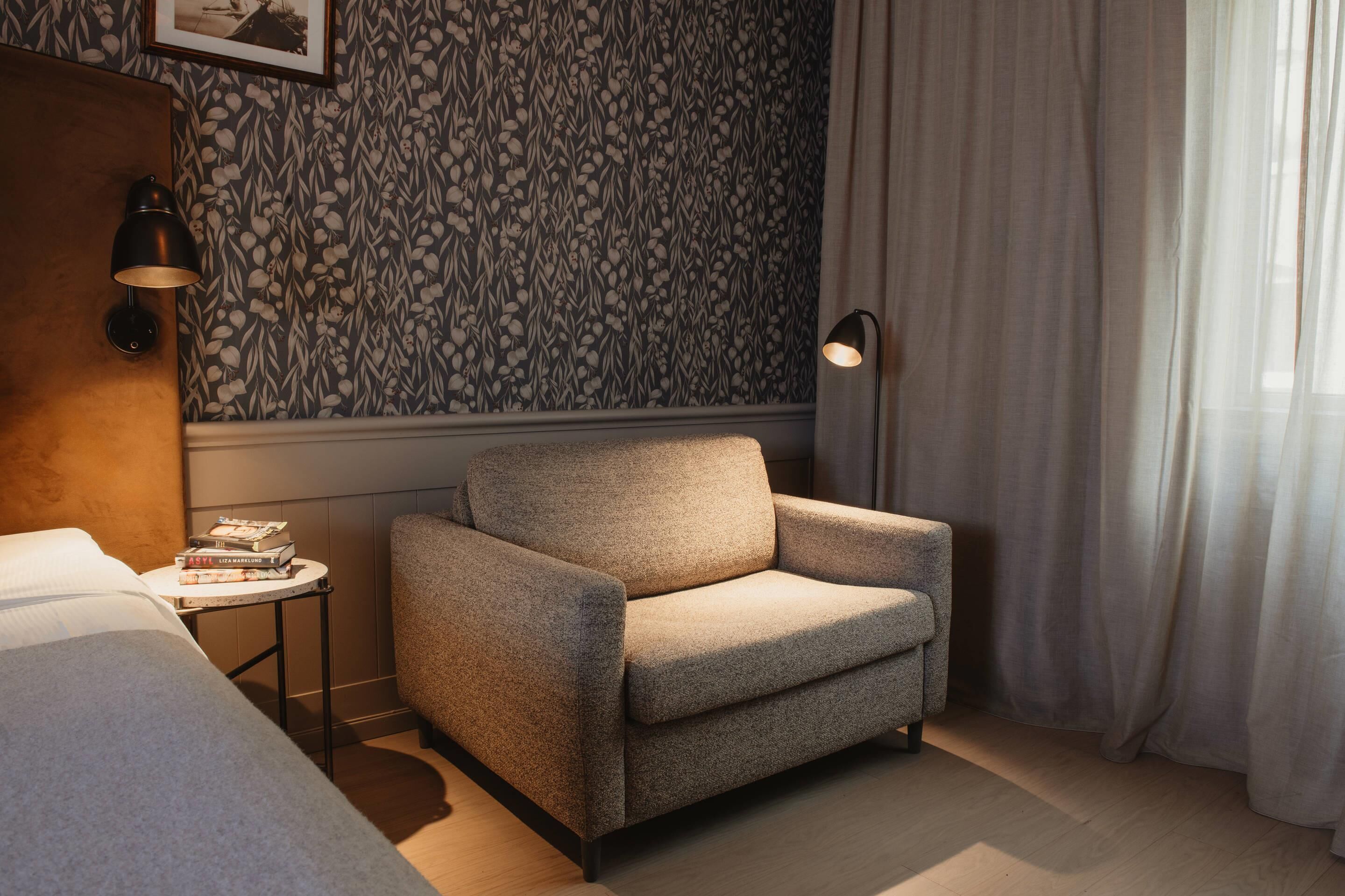 Quarto de hotel com papel de parede estampado, um sofá, uma mesa pequena com livros e uma cama.