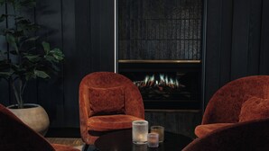A dark room with a fireplace, two red velvet chairs, a round table with candles, and a potted plant.