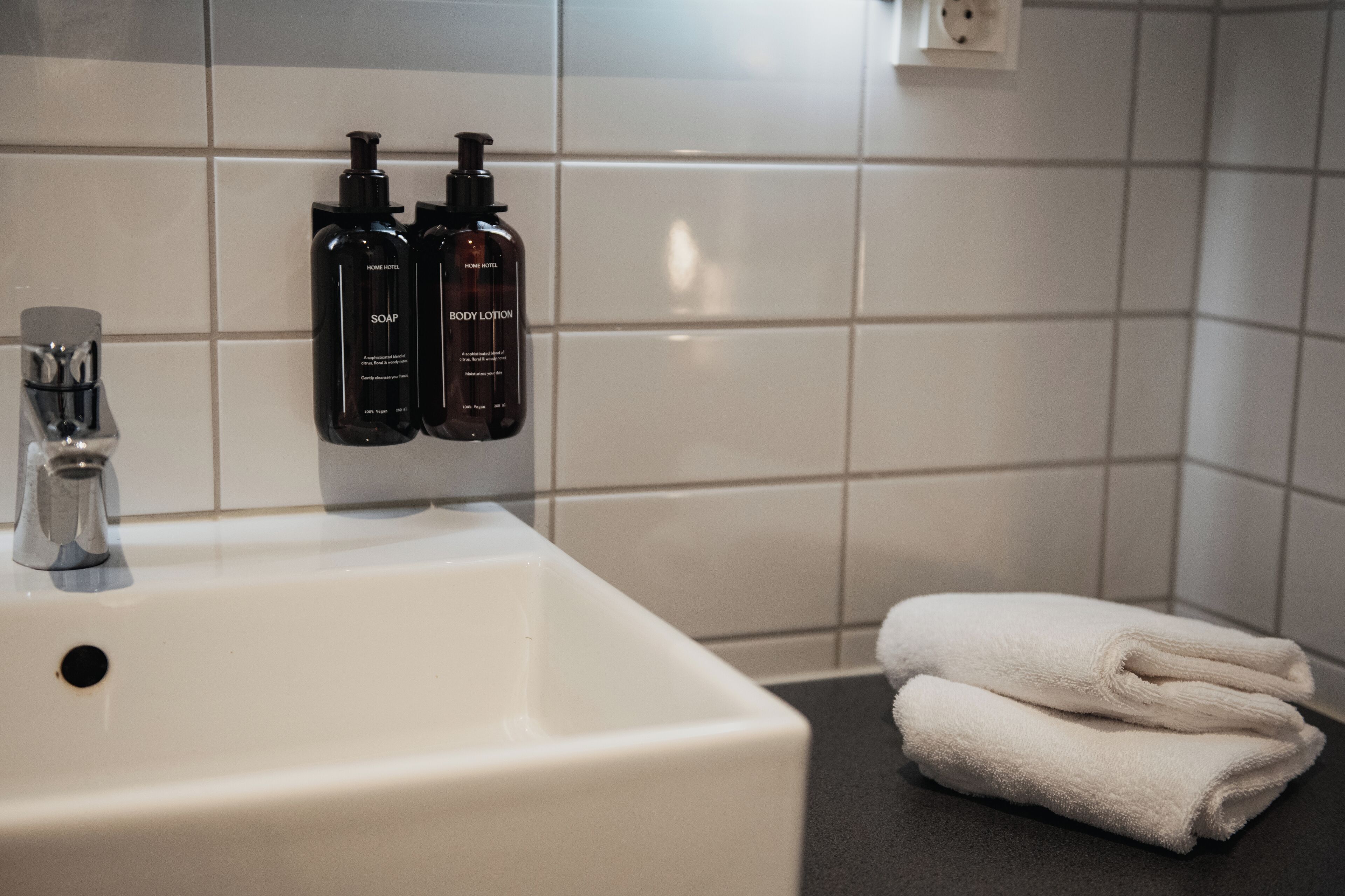Un baño con un lavamanos blanco, dos botellas de jabón y loción, y una pila de toallas blancas.