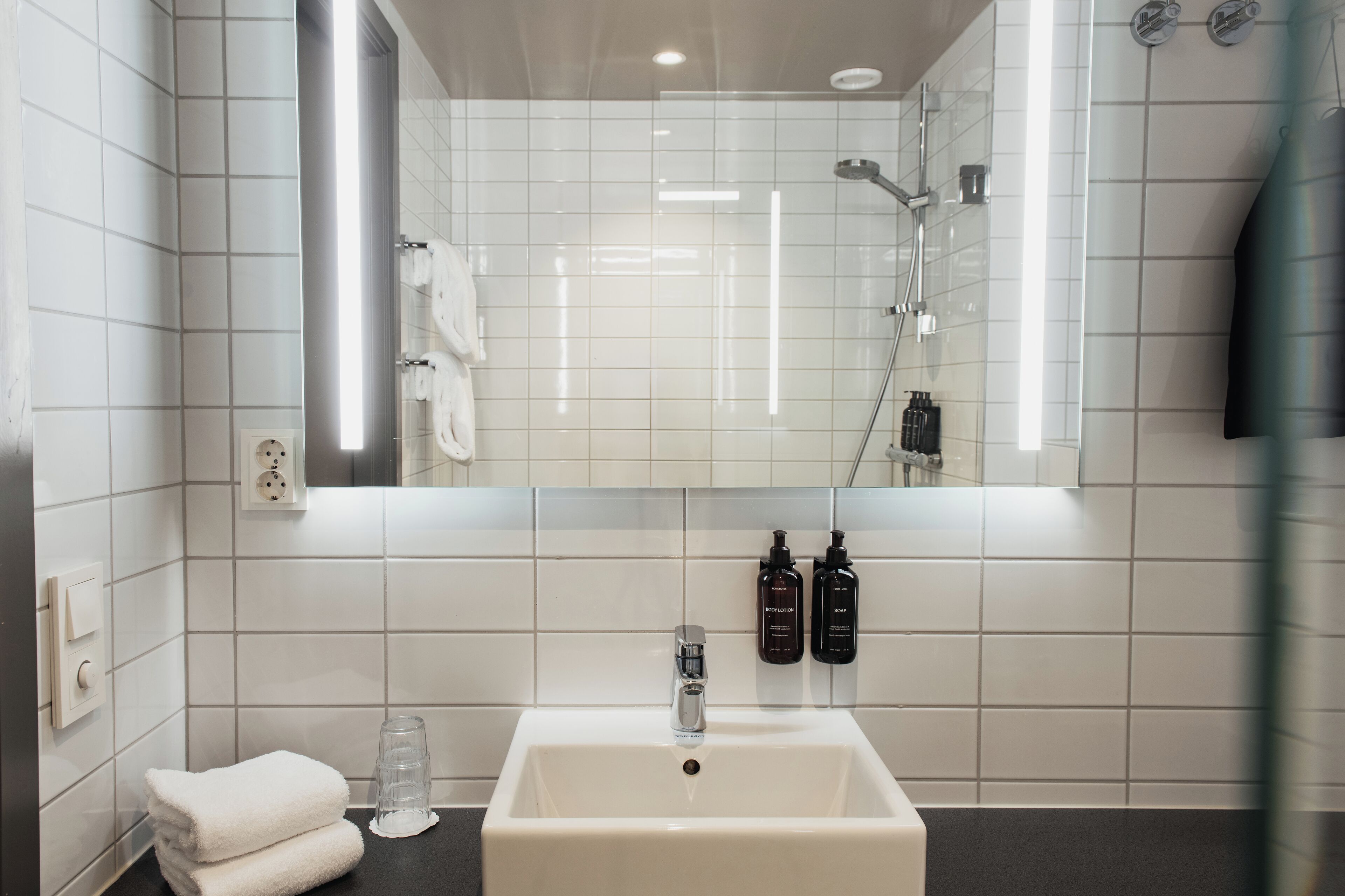 Une salle de bain avec des carreaux blancs, un lavabo, un miroir, un porte-serviettes et deux bouteilles de lotion pour le corps.