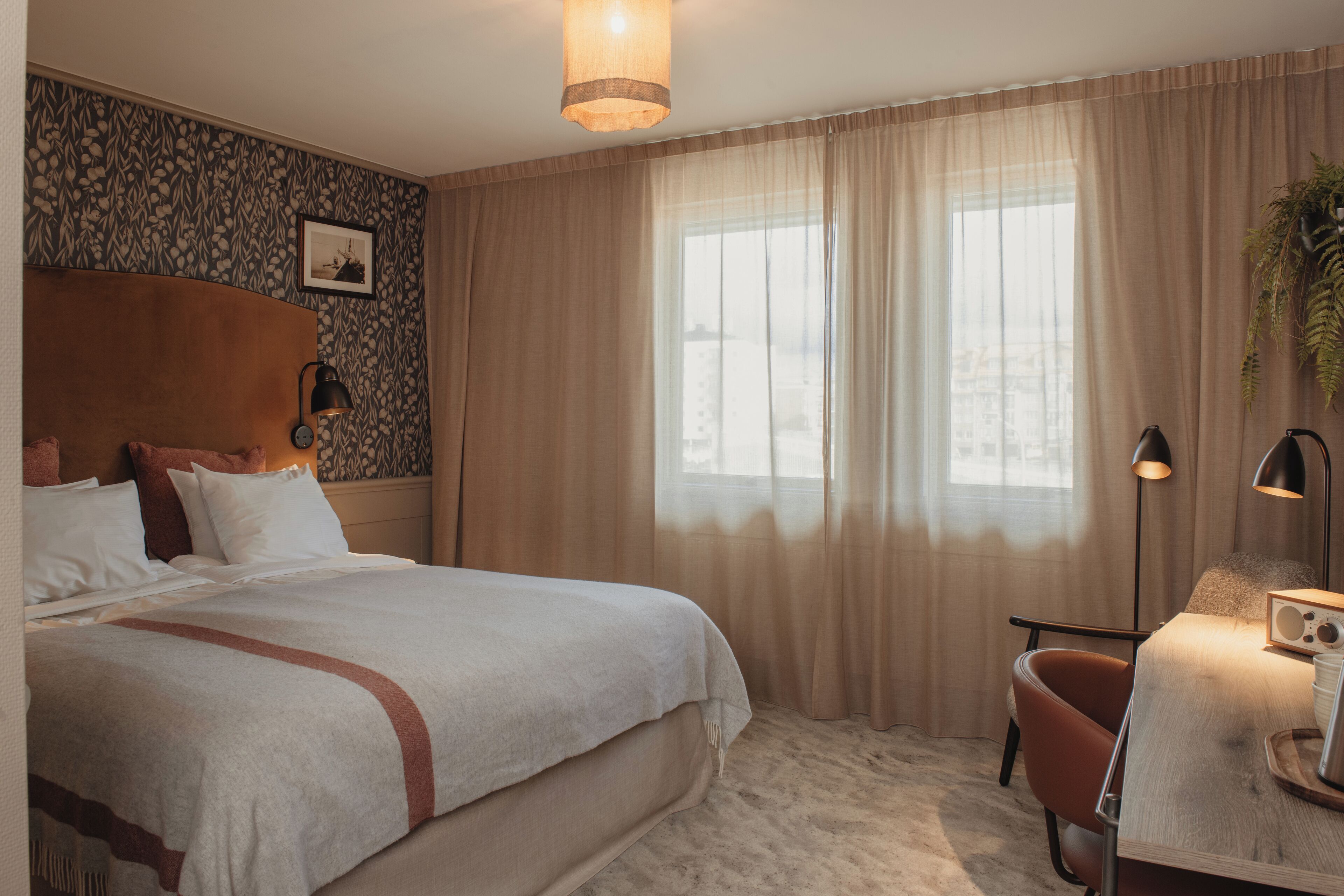 A hotel room with a large bed, a desk with a radio, a chair, and a window with curtains.