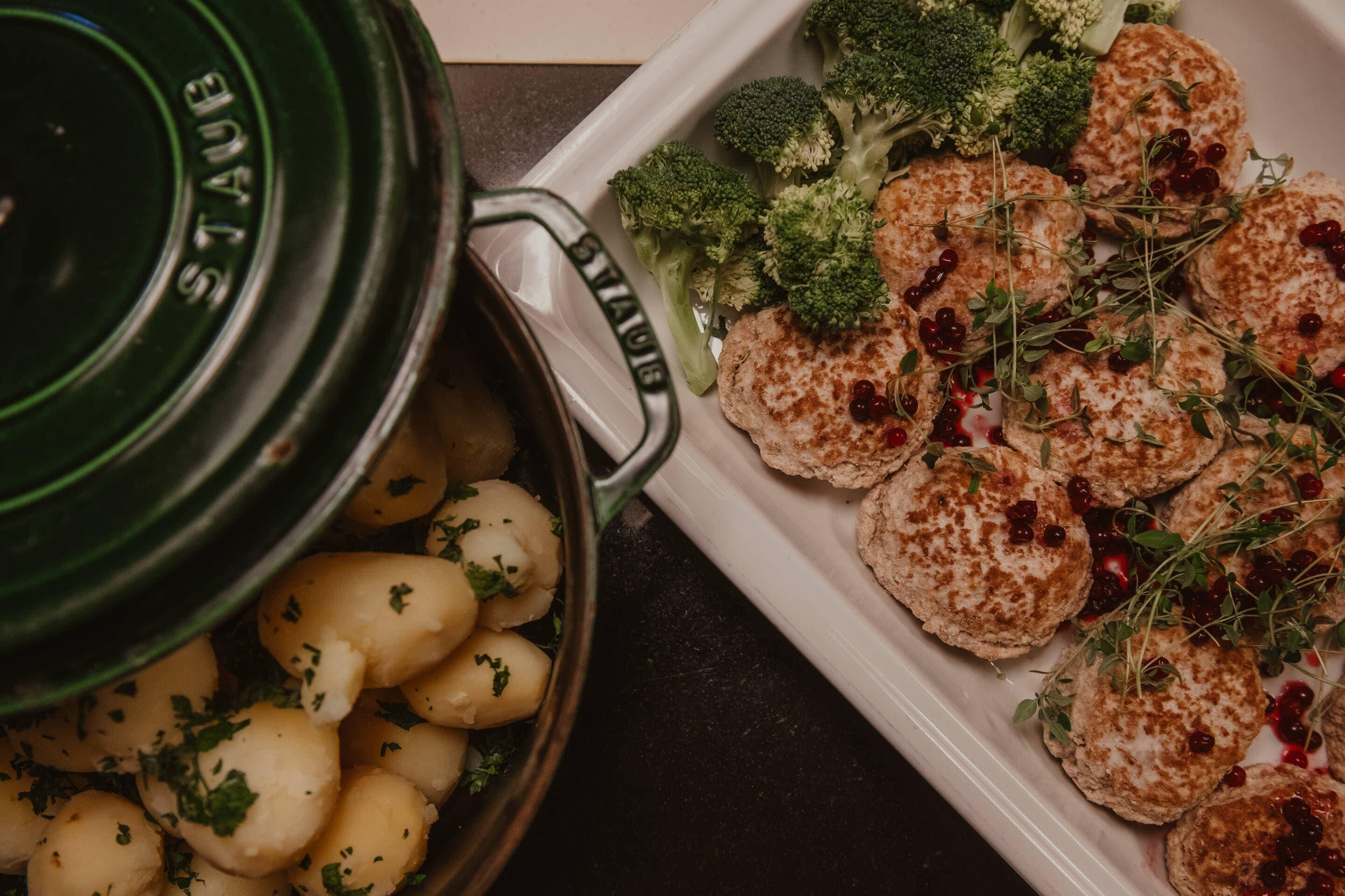 A pot of cooked potatoes with herbs, a white plate with meat patties and broccoli.