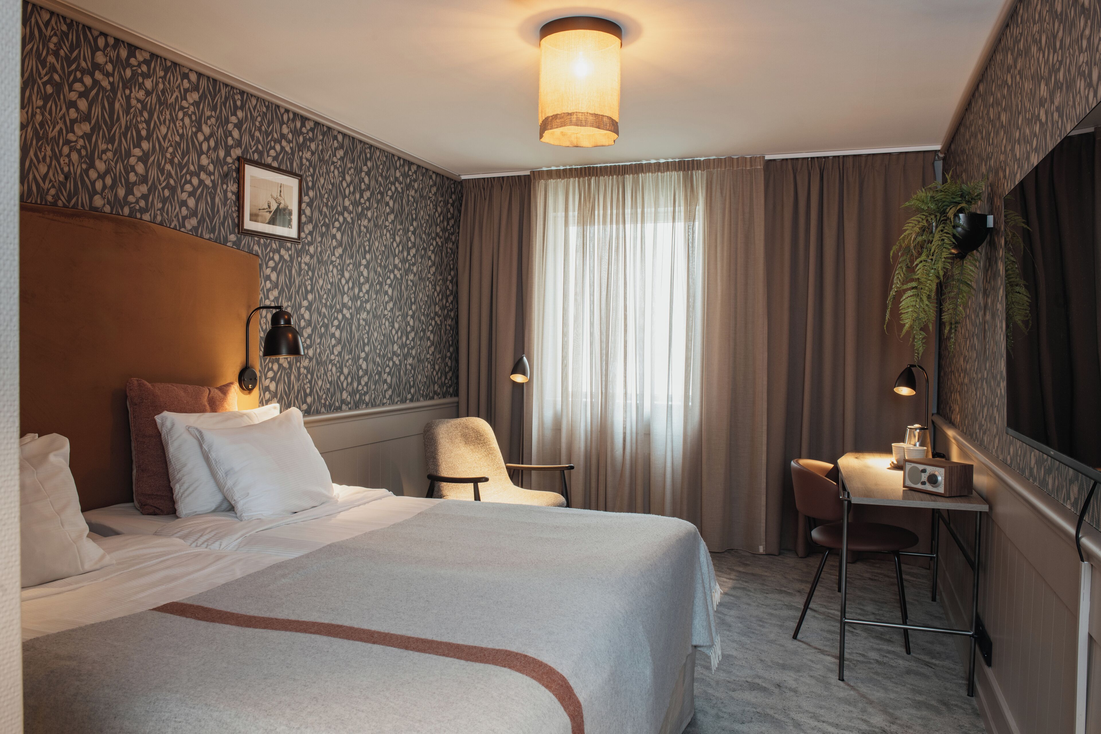 A hotel room with a large bed, a desk with a chair, a potted plant, and a large window with curtains.