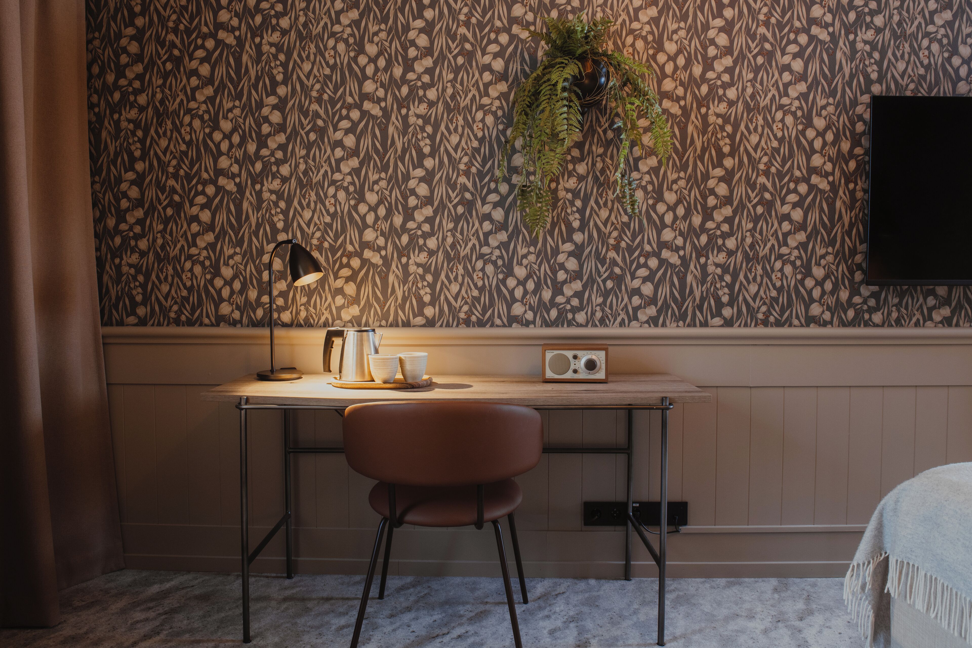 A room with a patterned wallpaper, a desk with a lamp, a chair, a radio, and a plant.