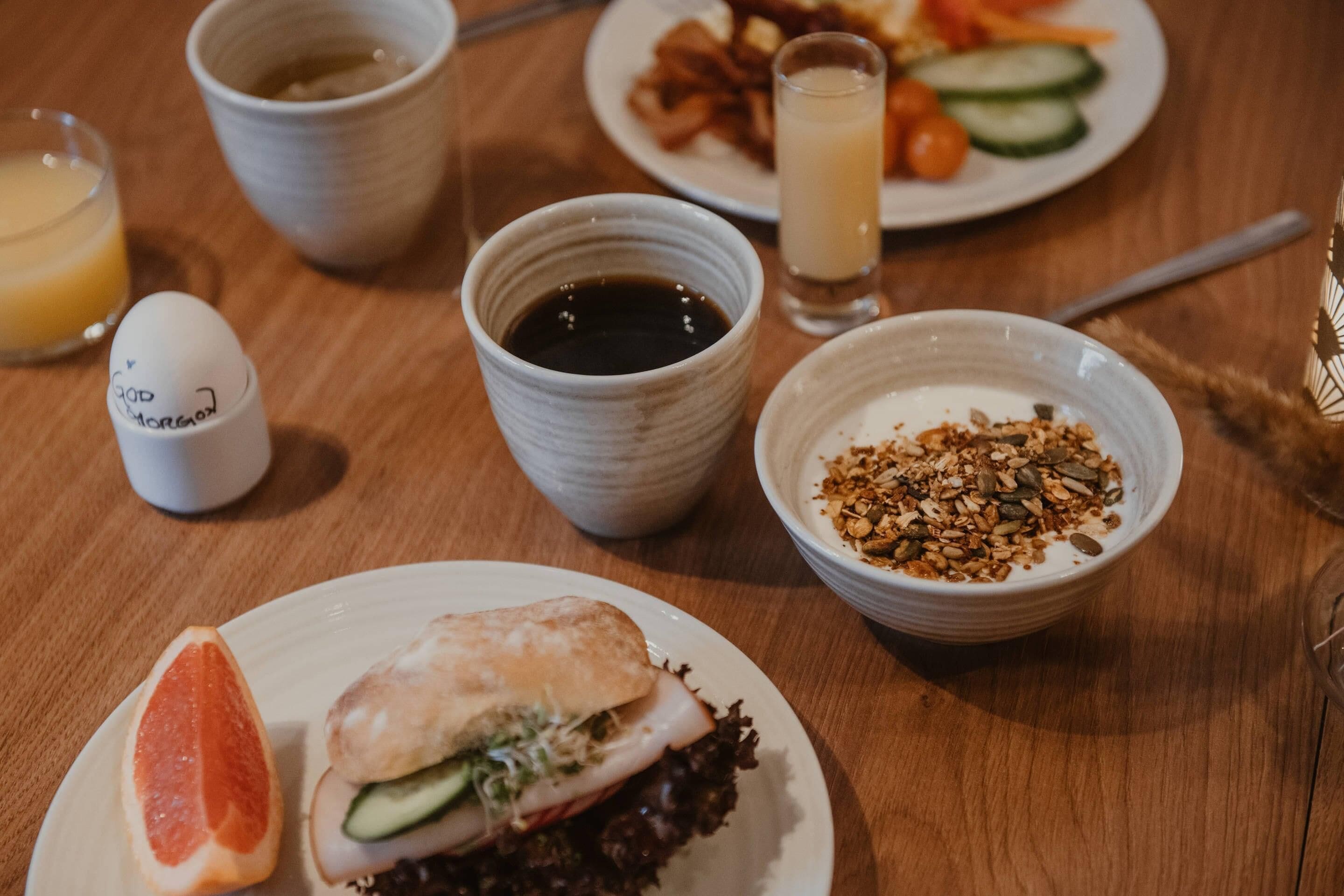 Mesa de café da manhã com pratos de comida, incluindo um sanduíche, um pote de granola e um copo de suco.