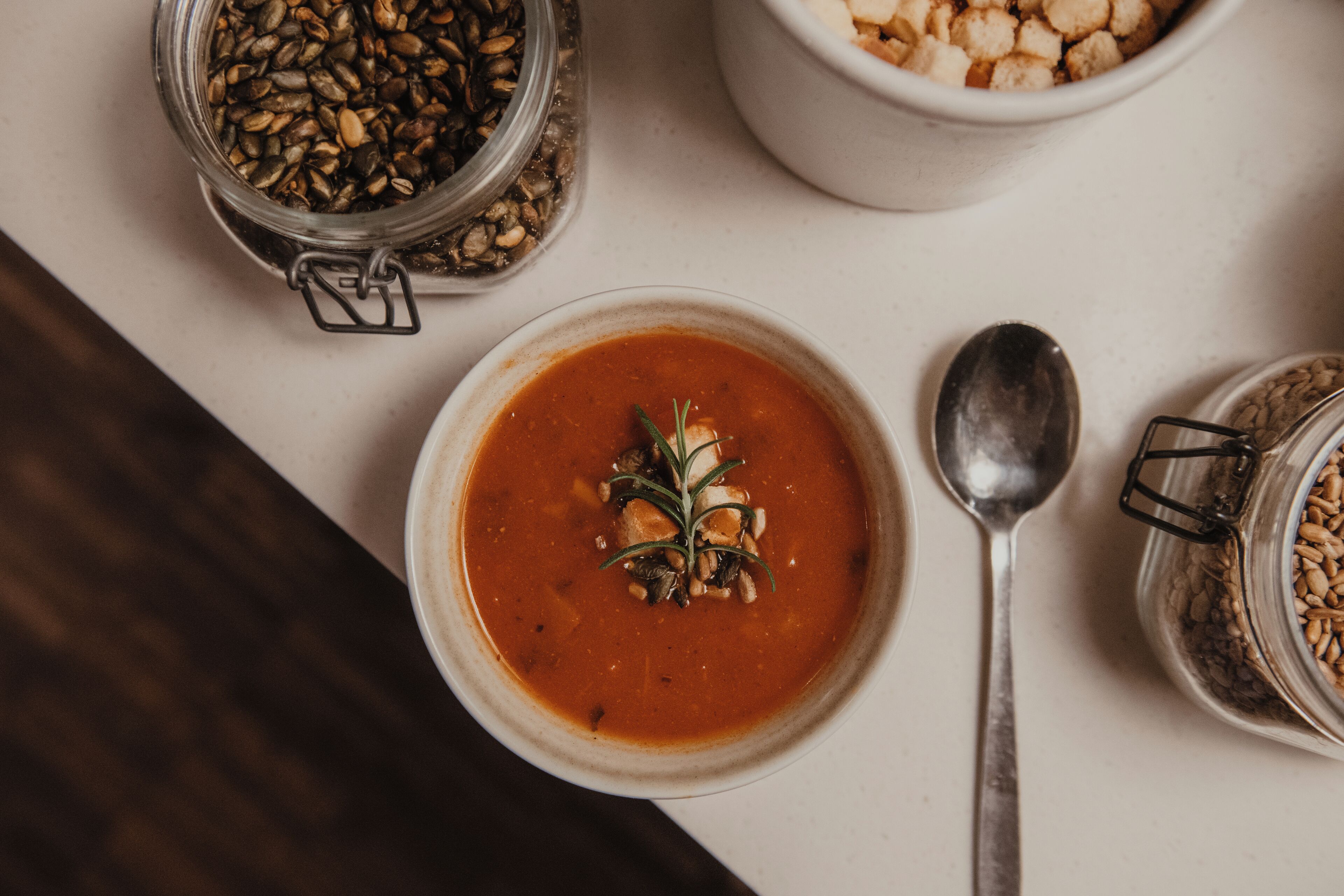 A bowl of soup with a garnish, a spoon, and two jars of seeds on a table.