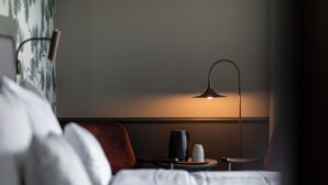 A hotel room with a bed, a bedside table with a lamp, a cup, and a vase.