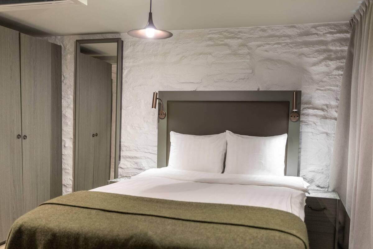 Una cama bien hecha con cabecera de madera, una lámpara colgante y una puerta que da a otra habitación.