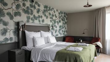 A neatly made bed with white linens, a green throw blanket, and towels on the side. There is a nightstand with a lamp and a small table with a cup and a book. The room has a tropical leaf wallpaper and a large window with curtains.