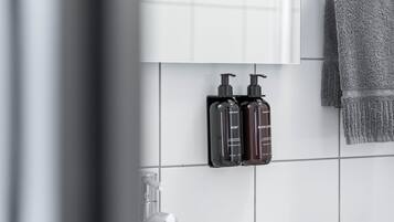 Casa de banho com parede revestida de azulejos brancos, dois dispensadores de sabonete pretos e uma toalha cinza pendurada num suporte.