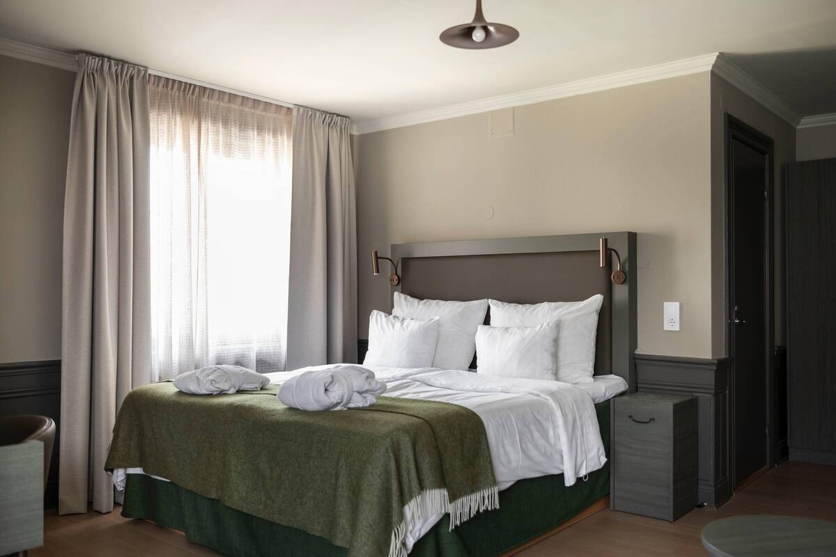 Una cama bien hecha con ropa blanca y un plaid verde, una mesita de noche y un pequeño mueble con cajones.