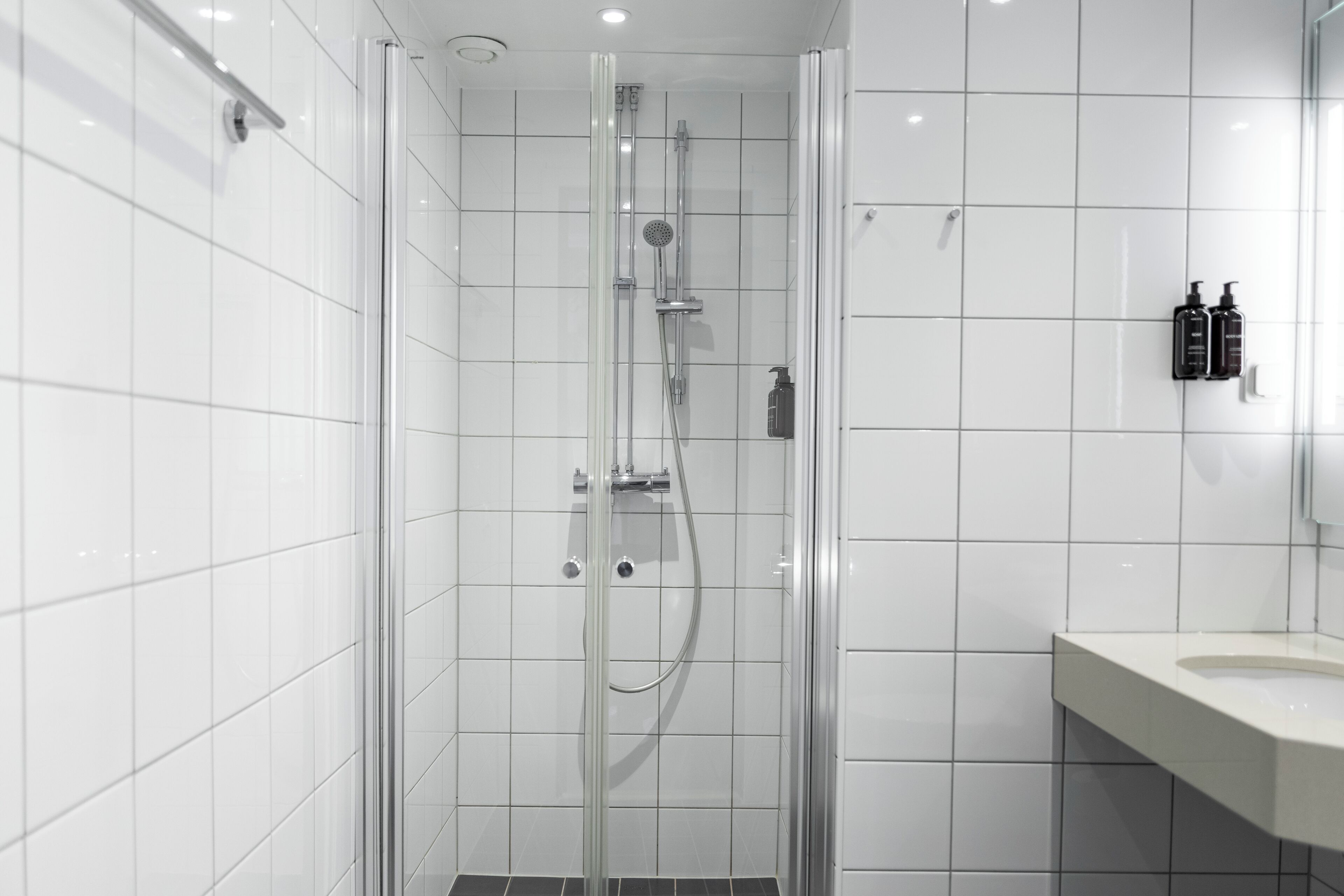 A modern bathroom with white tiles, a glass-enclosed shower, a sink, and two black toiletries on the wall.
